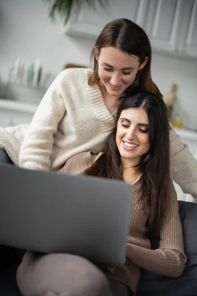 Femme brune utilisant un ordinateur portable flou près de petite amie en pull chaud à la maison — Photo de stock