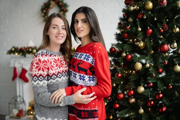 Lächelndes gleichgeschlechtliches Paar in Pullovern, das sich umarmt und in die Kamera am Weihnachtsbaum blickt — Stockfoto