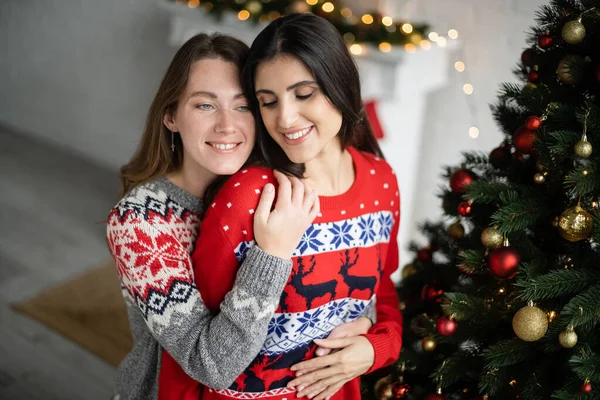 Улыбающаяся женщина в теплом свитере трогает партнера возле рождественской елки дома — стоковое фото