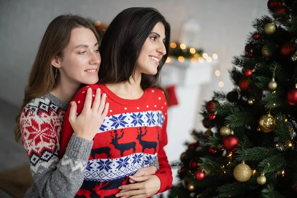 Mujer positiva abrazando pareja cerca del árbol de Navidad en la sala de estar - foto de stock