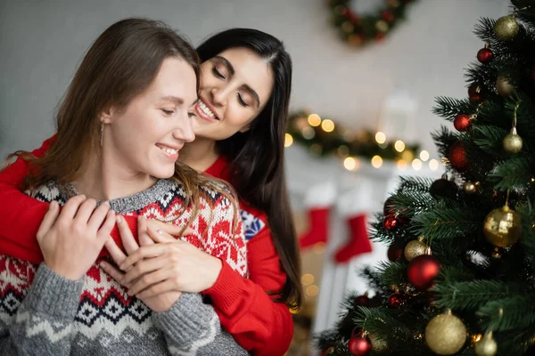 Улыбающаяся брюнетка обнимает подружку во время рождественского праздника дома — стоковое фото