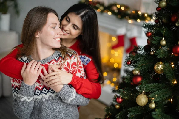 Young woman embracing girlfriend in sweater near decorated christmas tree — Stock Photo