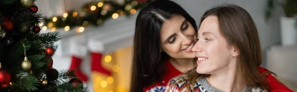 Усміхнена жінка цілує дівчину в светрі біля ялинки вдома, банер — стокове фото