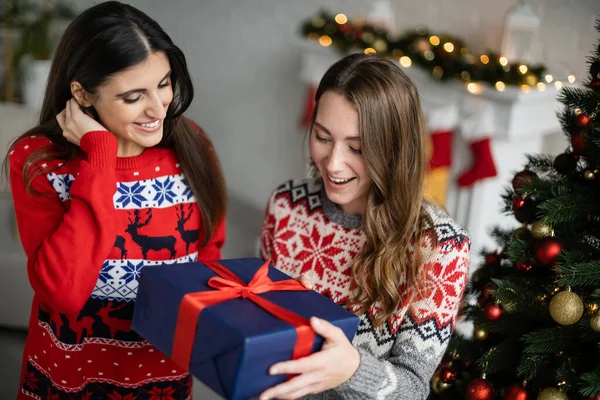 Femme gaie en pull tenant boîte cadeau près de petite amie et arbre de Noël à la maison — Photo de stock