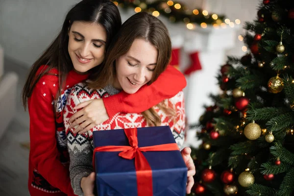 Позитивна жінка обіймає дівчину з подарунком біля ялинки вдома — стокове фото