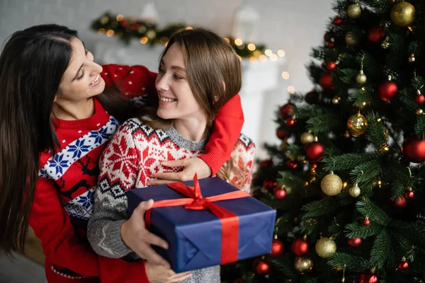 Positive woman hugging girlfriend with present during new year celebration in living room — Stock Photo