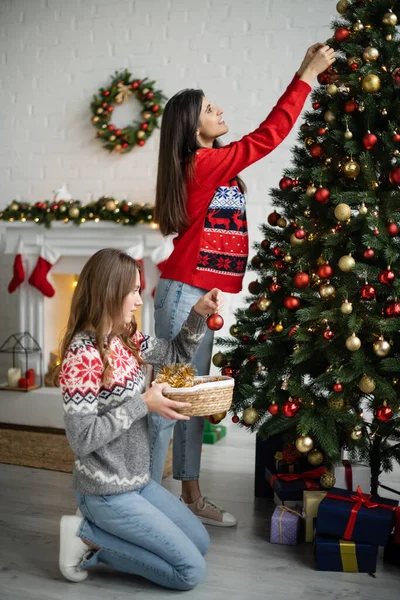 Vue latérale du couple lesbien décorant l'arbre de Noël près des cadeaux dans le salon — Photo de stock