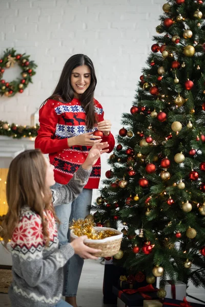 Mujer positiva sosteniendo la bola de Navidad cerca de novia y pino en la sala de estar - foto de stock