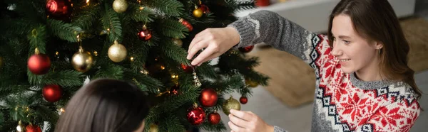 Vista ad alto angolo della donna sorridente in maglione che decora l'albero di Natale con la ragazza offuscata a casa, banner — Foto stock