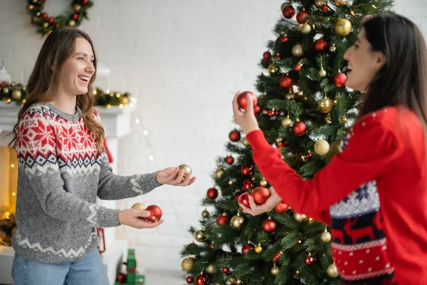 Positive same sex couple holding christmas balls near spruce tree in living room — Stock Photo