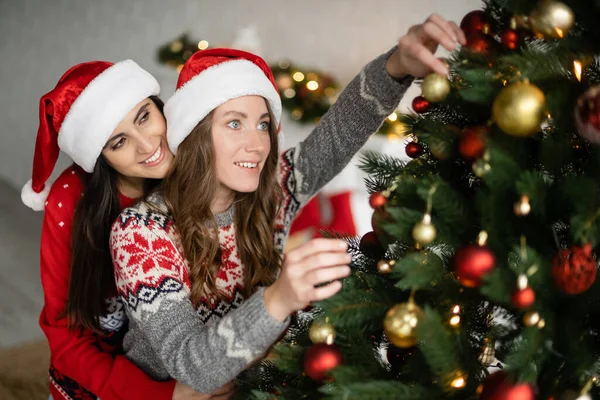 Позитивная лесбийская пара в шляпах Санты, украшающая рождественскую елку дома — стоковое фото