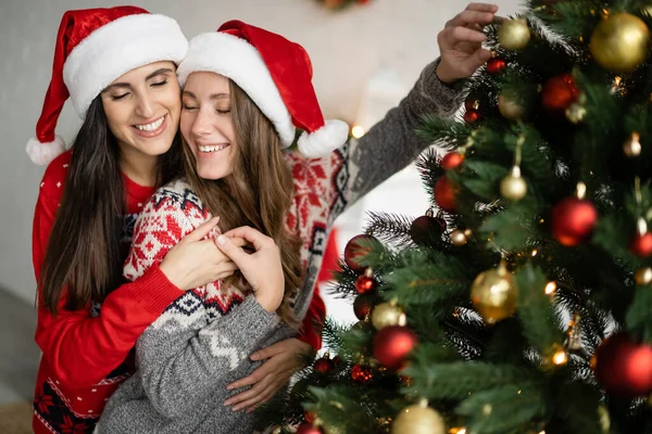 Positive same sex couple in santa hats hugging near blurred christmas tree at home — Stock Photo