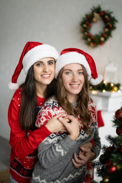 Cheerful same sex couple in santa hats hugging and looking at camera near christmas tree at home — Stock Photo