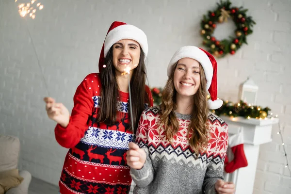 Happy lesbian couple in santa hats holding sparklers in living room — Stock Photo