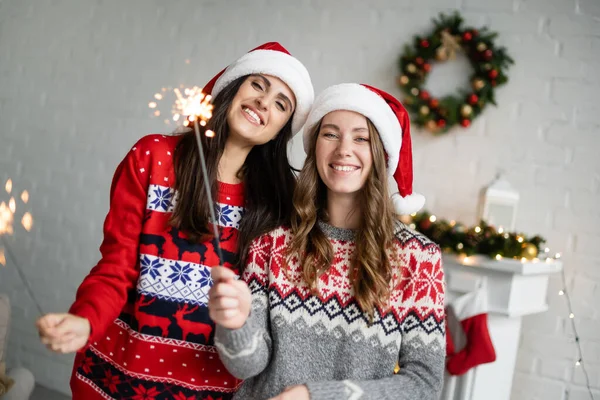 Positive lesbian couple in sweaters and santa hats holding sparklers in living room — Stock Photo
