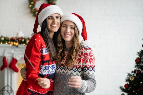 Happy same sex couple in santa hats holding sparklers and looking at camera at home — Stock Photo