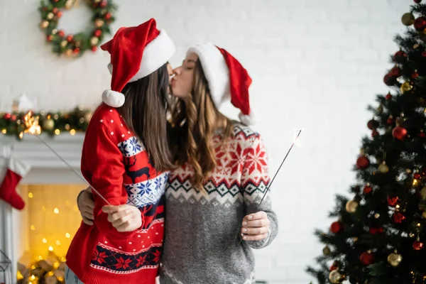 Side view of blurred lesbian couple in santa hats kissing and holding sparklers at home — Stock Photo