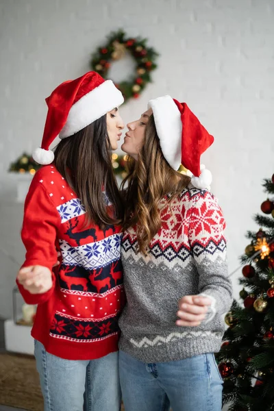 Seitenansicht eines gleichgeschlechtlichen Paares mit Weihnachtsmannhüten, die sich küssen und Wunderkerzen im Wohnzimmer halten — Stockfoto