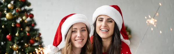 Веселая лесбийская пара в шляпах Санты, смотрящая в камеру возле искорки и рождественской елки дома, баннер — стоковое фото