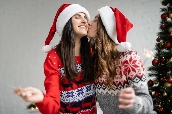 Junge lesbische Frau in Pullover und Weihnachtsmütze küsst Freundin mit Wunderkerze zu Hause — Stockfoto