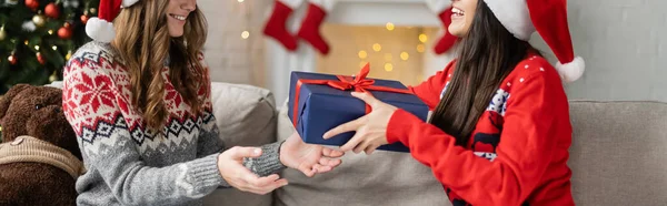 Vue recadrée de femme lesbienne gaie dans le chapeau de Père Noël et le pull donnant cadeau à la petite amie à la maison, bannière — Photo de stock