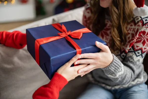 Vista recortada de la mujer en suéter dando regalo de Navidad a la novia en el sofá en casa - foto de stock