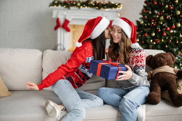 Lesbian woman in santa hat kissing girlfriend with gift box on couch in living room — Stock Photo