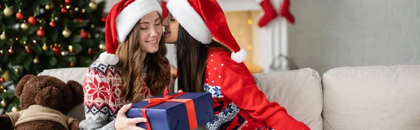 Позитивная женщина в шляпе Санты целует подругу с подарком во время празднования Рождества дома, баннер — стоковое фото
