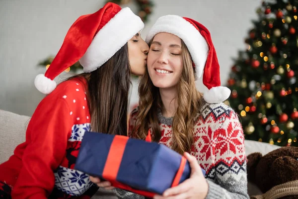 Lesbian woman in sweater kissing girlfriend in santa hat holding gift at home — Stock Photo