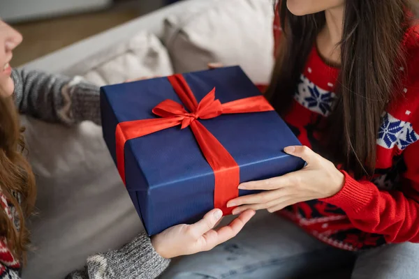 Vista recortada de la mujer joven dando caja de regalo a la novia en suéter de Navidad en casa - foto de stock