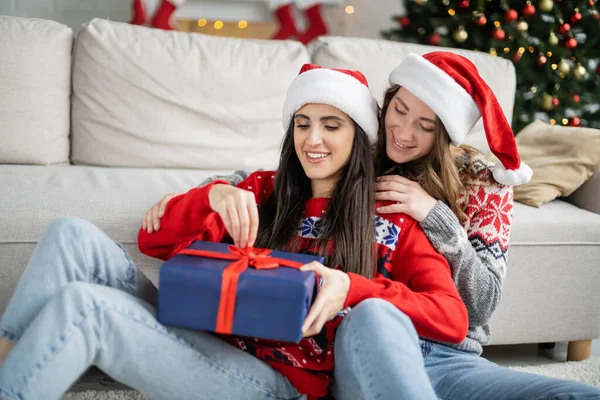 Donna sorridente in cappello di Babbo Natale abbracciando fidanzata con regalo di Natale in soggiorno — Foto stock