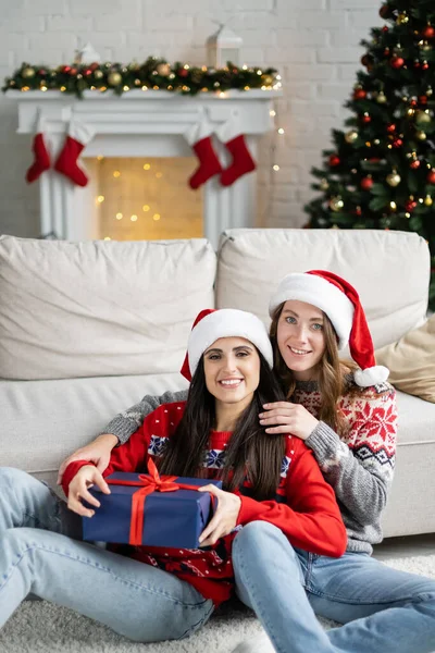 Positive same sex couple in santa hats holding new year gift near couch at home — Stock Photo