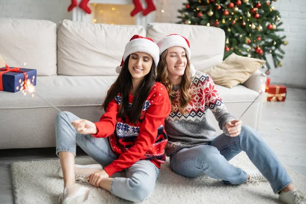 Junges lesbisches Paar mit Weihnachtsmann-Hüten und Wunderkerzen zu Hause — Stockfoto