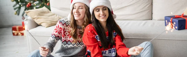 Jeune couple de même sexe dans des chapeaux Santa tenant des étincelles dans le salon, bannière — Photo de stock