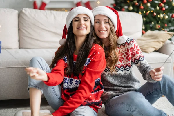 Positive lesbian couple in santa hats holding sparklers in living room — Stock Photo