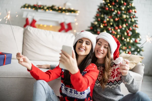 Cheerful same sex couple in santa hats taking selfie on smartphone and holding sparklers at home — Stock Photo