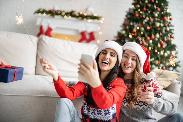 Позитивная лесбийская пара в шляпах Санты с помощью смартфона и с искрами в гостиной — стоковое фото