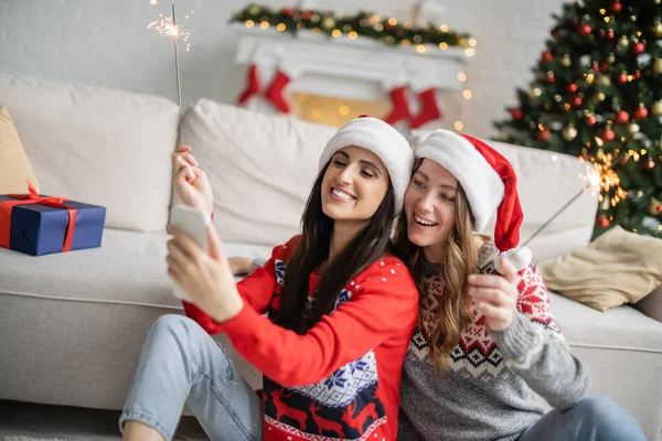 Positive lesbian couple using smartphone and holding sparklers during new year celebration at home — Stock Photo