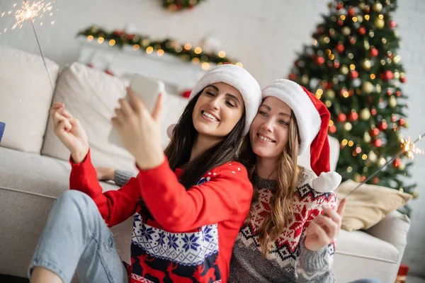 Alegre lesbiana pareja en santa sombreros tomando selfie y celebración sparklers en sala de estar - foto de stock