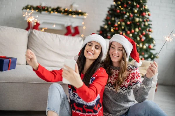 Positive same sex couple using smartphone and holding sparklers during new year at home — Stock Photo