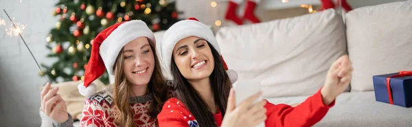Alegre lésbicas casal no santa chapéus e camisolas usando smartphone e segurando sparklers em casa, banner — Fotografia de Stock