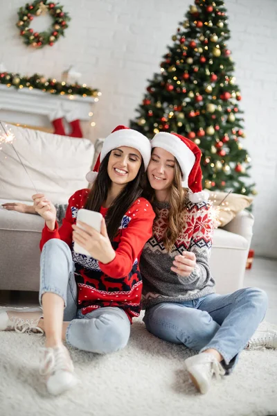 Молодая лесбийская пара в шляпах Санта-Клауса пользуется мобильным телефоном и держит бенгальские огни во время Рождества — стоковое фото