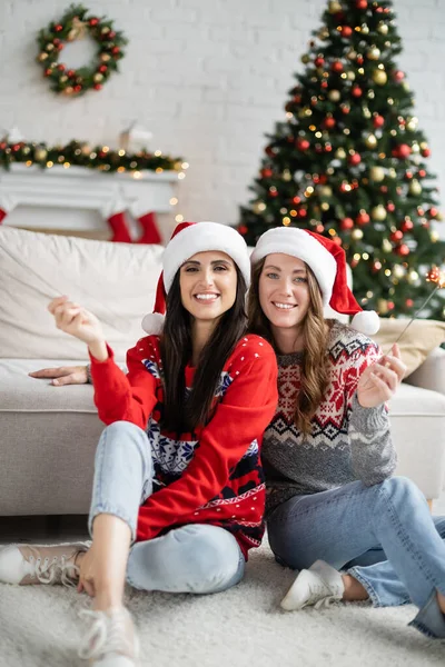 Cheerful lesbian couple in santa hats holding sparklers during new year celebration at home — Stock Photo