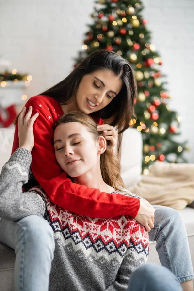 Cheerful young woman touching hair of girlfriend in sweater at home — Stock Photo