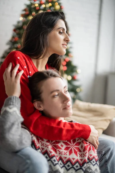 Joven lesbiana mujer abrazando novia en navidad suéter en casa - foto de stock