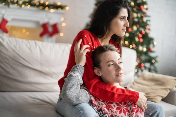Couple de même sexe dans les chandails de Noël étreignant sur le canapé à la maison — Photo de stock