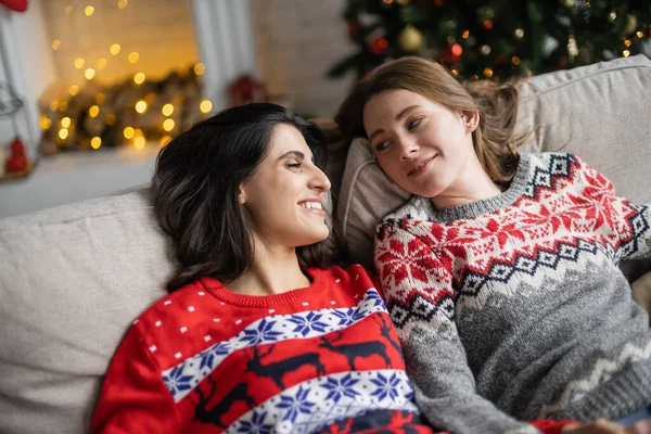 Позитивная лесбийская пара в новогодних свитерах отдыхает дома на диване — стоковое фото