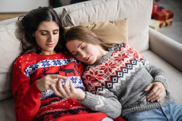 Young same sex couple in christmas sweaters touching blurred hands on couch at home — Stock Photo