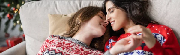 Лесбийская пара в рождественских свитерах, касающихся рук и целующихся дома, баннер — стоковое фото