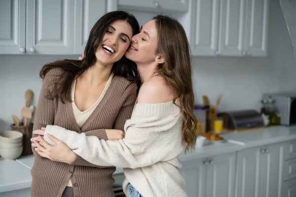 Positive woman in sweater hugging girlfriend in kitchen — Stock Photo